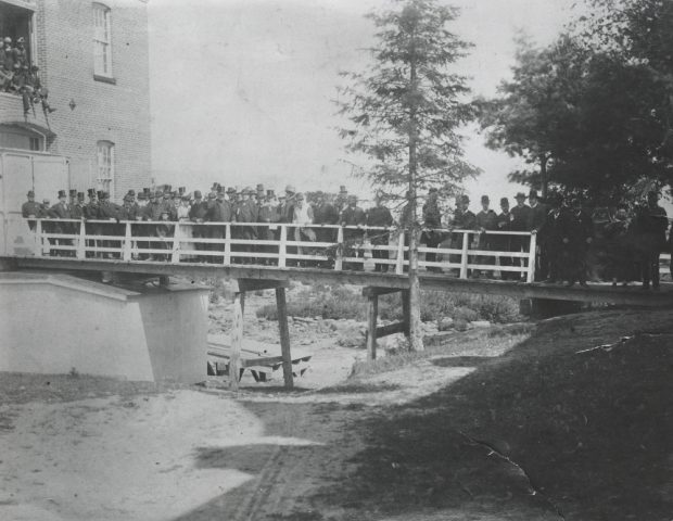 Black & white photograph depicting several men and women standing on a bridge. Part of the Saint-Jérôme paper mill is seen at left, along with several trees at right.