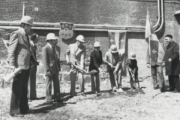 Black & white photograph depicting several men standing together, wearing suits and hardhats and holding shovels.