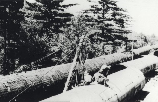 Black & white photograph showing workers next to large water pipes in a woodland.