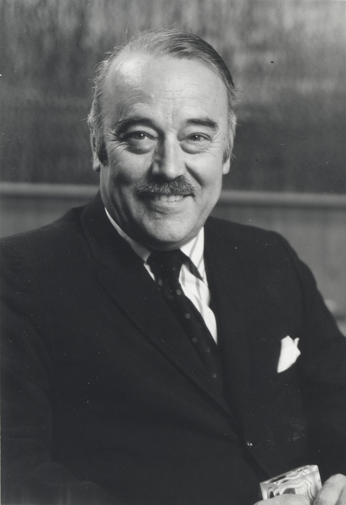 Black & white portrait of a smiling man in his forties or fifties, with a small moustache; he wears a dark suit and tie.