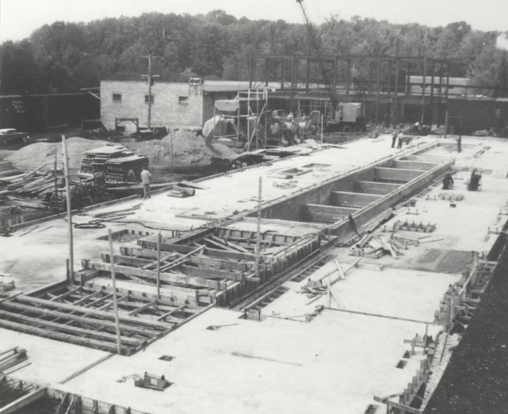 Photographie noir et blanc d’un bâtiment en construction. S’y trouvent des ouvriers et de la machinerie. 