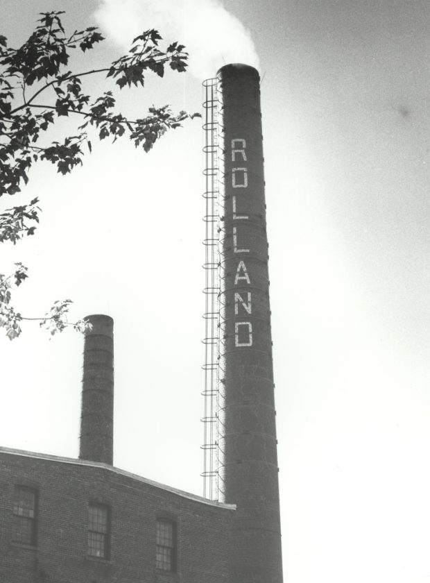 Black & white photograph of a factory smokestack bearing the name Rolland.