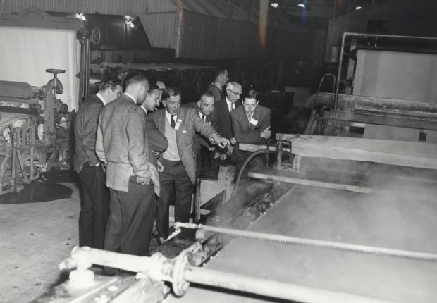 Black & white photograph depicting a group of men beside a paper machine. In the background, another machine is seen, at left, along with the interior of the plant.