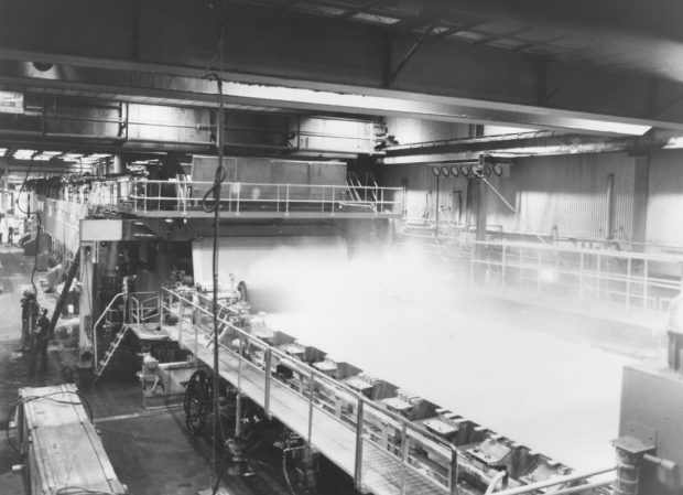 Black & white photograph of a working paper machine. Vapour escapes from one of the trays, visible in the foreground. On the left are workers close to the machine.
