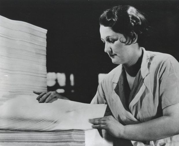 Black & white photograph depicting a woman counting a stack of sheets.