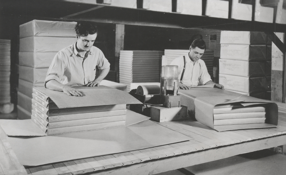 Photographie noir et blanc représentant deux hommes en train d’emballer des paquets de feuilles. En  arrière-plan se trouvent d’autres paquets emballés. 