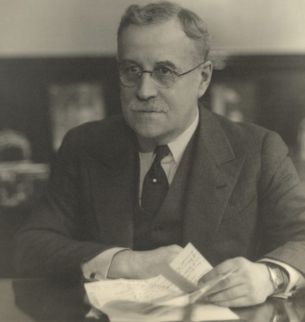 Black & white portrait of a man wearing glasses and a suit, seated at a desk and holding a sheet of paper.