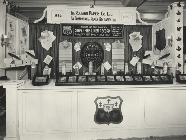 Black & white photograph of a stand displaying numerous photographs, paper samples and glass containers. Posters advertise the Rolland Company name.