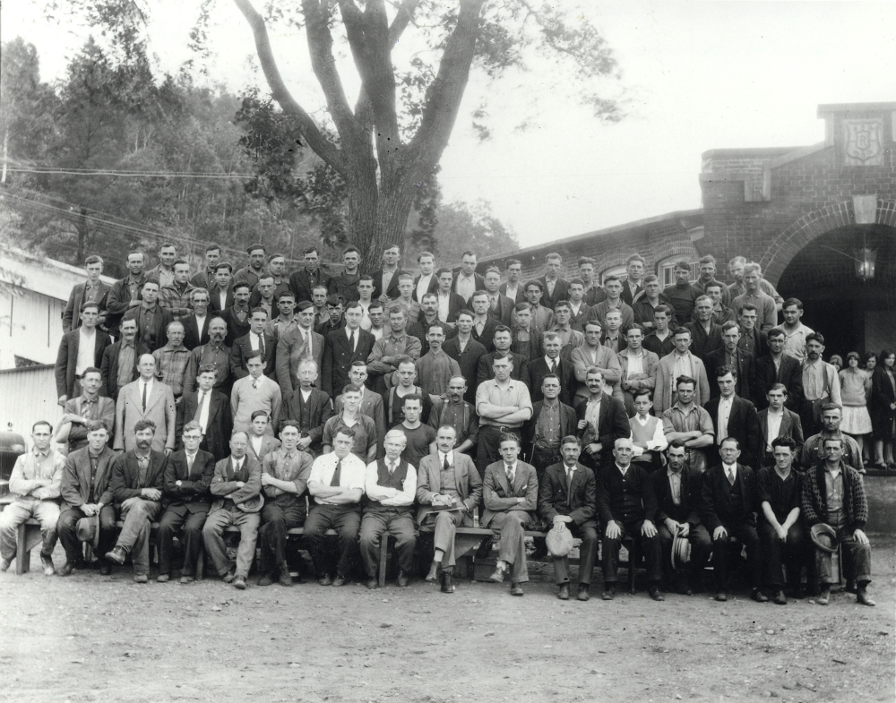 Black & white photograph large group of men standing in rows in front of a factory.