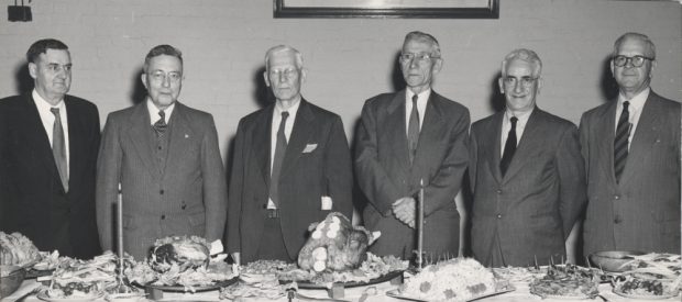Black & white photograph of six men standing behind a table.