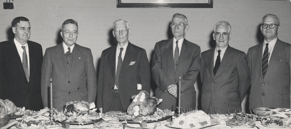 Black & white photograph of six men standing behind a table.