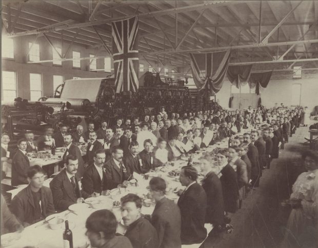 Black & white photograph showing several people seated at long tables in a factory setting. A paper-making machine stands in the background.