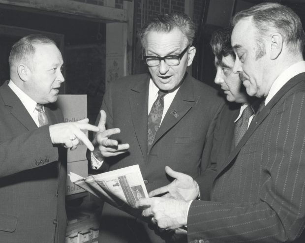 Black & white photograph of a group of men looking at a newspaper; one appears to be talking.