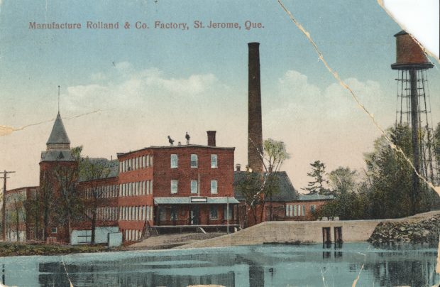 Colour image of buildings close to a large chimney stack and a river.