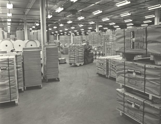 Black & white photograph of a huge room containing many stacked packages and rolls.