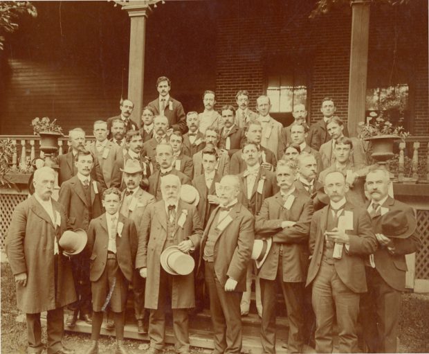 Sepia-tone photograph showing a group of men clad in suits, on stairs in front of a house.