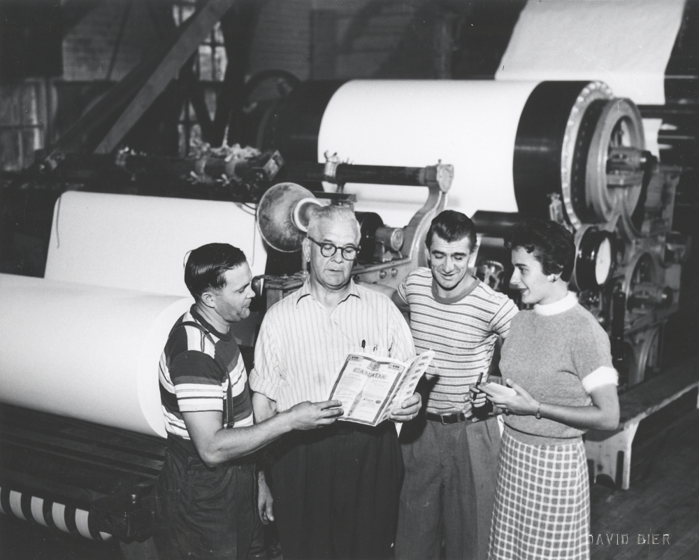 Photographie noir et blanc illustrant quatre personnes devant une machine à papier. 
