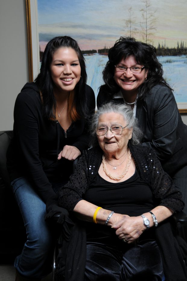A woman seated in wheelchair with two women behind her. On the wall behind them there is a painting of snow and trees.