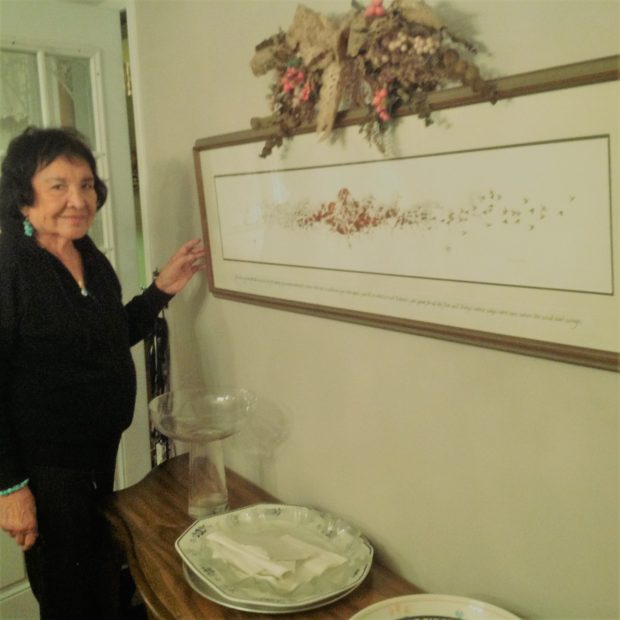 An elderly Lina Gallup standing beside a table and photo on the wall.