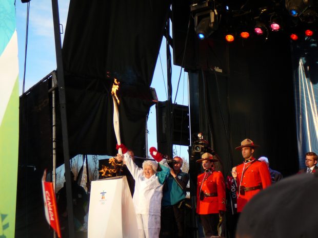 Elder Elsie Yanik on stage at the Olympic Torch Relay as a torch bearer, wearing an authentic Olympic tracksuit with red mittens with Canadian maple leaves on them, holding a white torch. Two mounted police stand behind her in red uniforms.