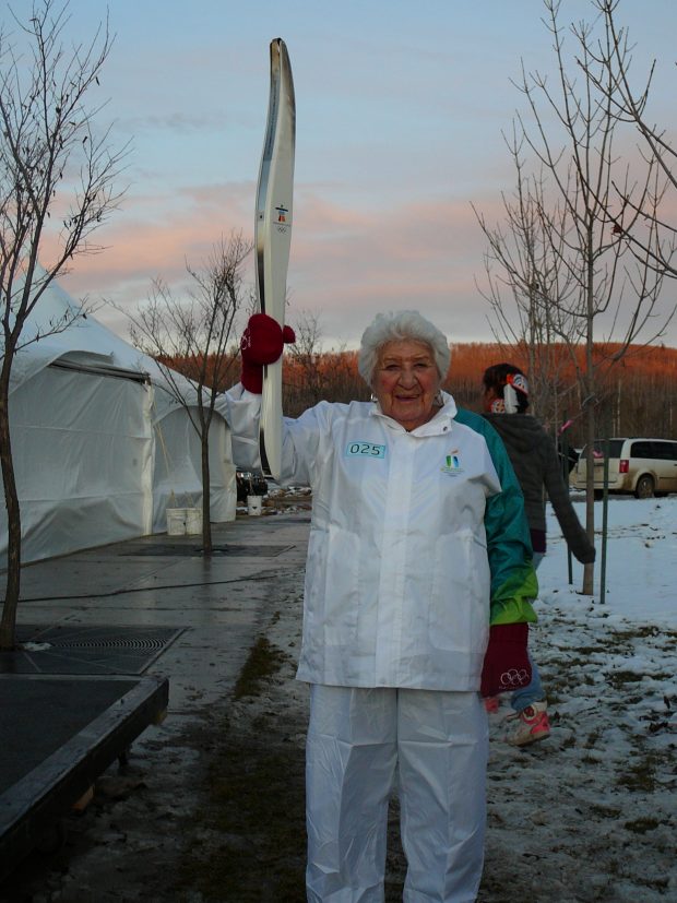 An elderly Elsie Yanik holding a white and silver Olympic torch on a winter’s day, a woman walks behind her along with tents and trees.