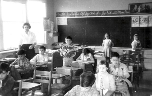 A black and white photo of Mrs. Golosky’s classroom at St. John’s School in the late 1960s, students sit in wooden desks facing the front, a young girl is shown in the back by a large chalkboard.