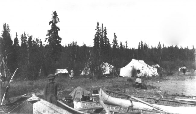 A historic photo from the 1920s of camp set up with over four tents, canoes sit alongside the river with children around them.