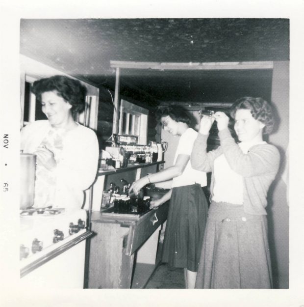 A middle-aged Nancy Woodward stands in the kitchen stirring a pot of jam with two other women in the background, one is fixing something on a second stovetop.