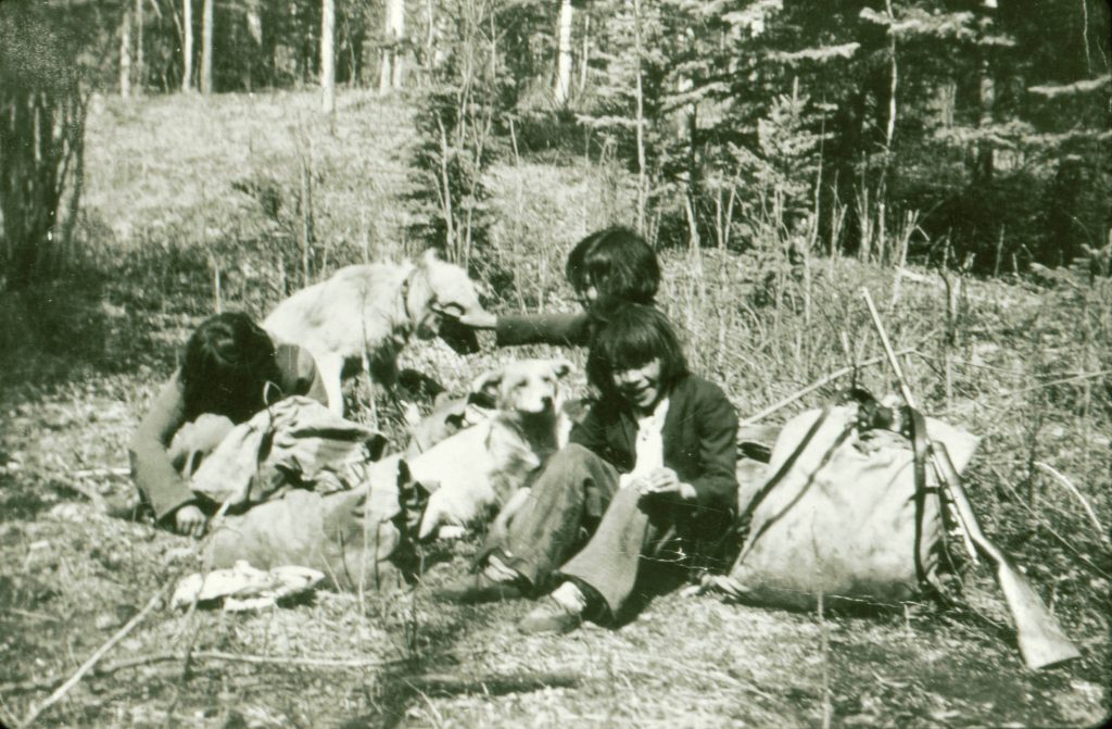 Gerty Sanderson as a young girl sits on a grassy forest floor beside a large sack and rifle with her two younger sisters next to two dogs, one is petting one of them.