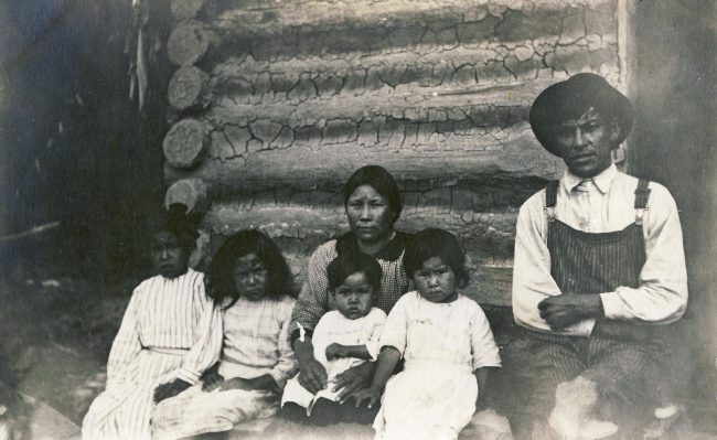 A black and white photo of an Indigenous family with four children sitting in front of a log cabin.