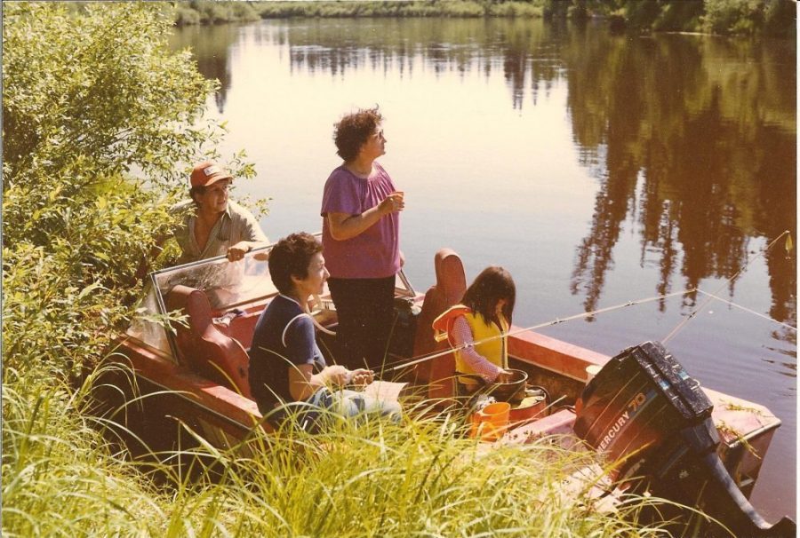 A coloured photo from the 1980s of people in a boat on the river, the forest trees are reflected in the water.
