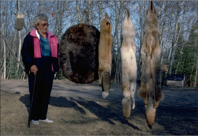 An elderly Katie Sanderson stands alongside four different furs, one round and brown, the three others are fox furs, varying in size.
