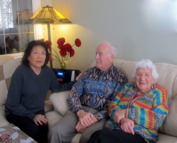 An elderly Elsie Yanik sits on a couch wearing a colourful patterned shirt beside an elderly man and woman, red flowers are shown behind them on a lamp table.
