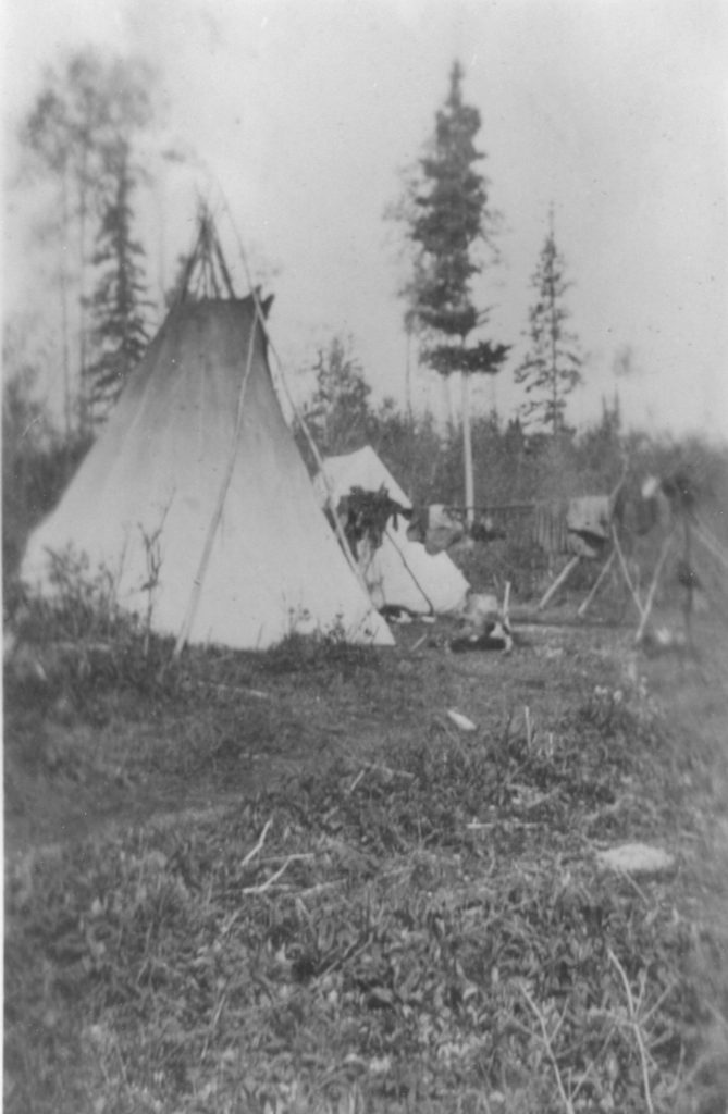 A black and white photo of two teepees sitting next to each other with trees in the background, one standing taller than the other.