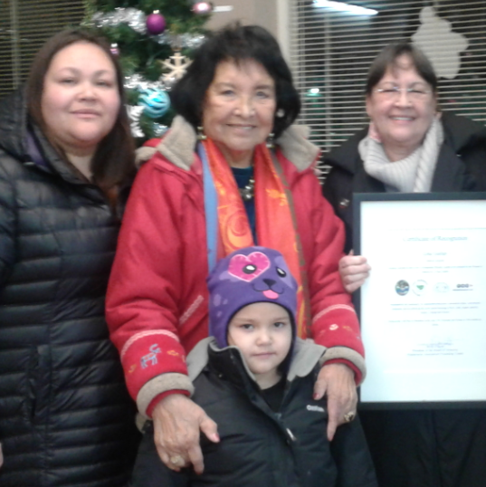 A group of women standing close together Lina Gallup in the middle of her grandson’s wife on the right and her daughter Cecile Calliou on the right side, holding a certificate and the great-granddaughter standing in front of Lina all women wearing their winter coat standing behind a Christmas tree.