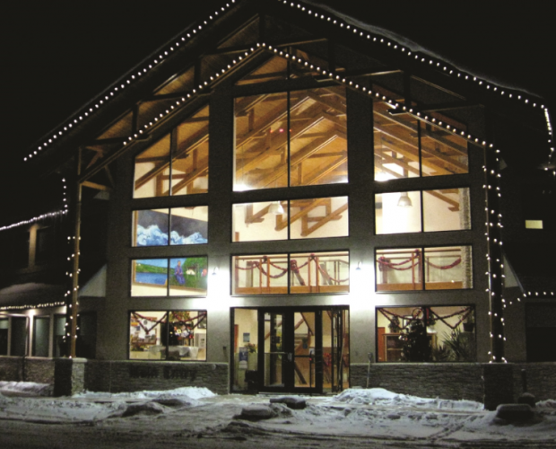 A photo of a modern structure of a building with exposed big trusses and large big windows that make the inside of the building visible from the outside. White lights decorated around the front frame of the roof.