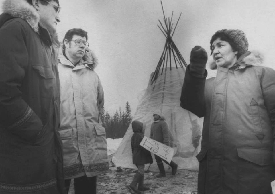 Black and white photo of two men and a woman standing close to each other, the woman is Dorothy McDonald shown pointing and as if speaking toward the men with a teepee standing in the background and a young child holds a sign board and some trees on the far distance.