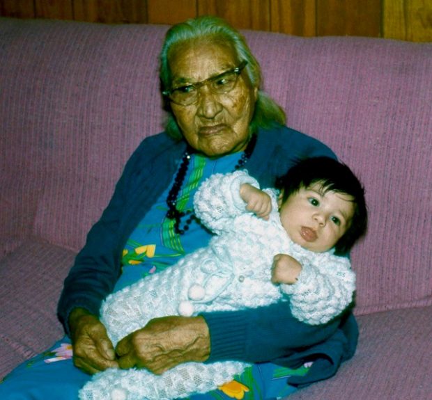 An elderly Mary-Rose sits on a pink sofa wearing a blue cardigan and black beaded necklace with glasses on, holding her great-great-grandson dressed in a white onesie.