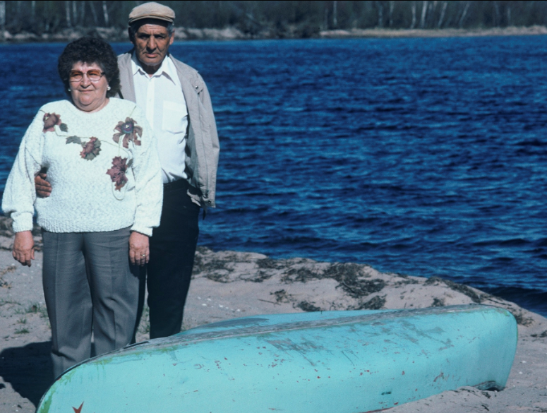 An elderly Nancy and Bill Woodward stand beside a light blue canoe on a sandy beach, ripples of waves are shown on the lake in the background.