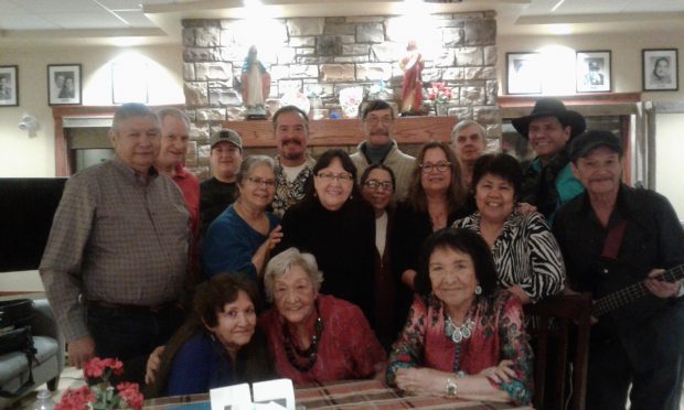 An elderly Lina Gallup sits at a table with over a dozen of men and women surrounding her with two other elderly women sitting and the others standing inside a family home with a large stone fireplace in the background.