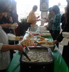 A group of people around a table with different types of food, soup, bannock, fruits along both side of the table with a green table plastic cloth.