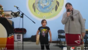 A coloured picture with a medicine wheel on the background on a stage an Elderly woman and a young girl. The elderly woman is holding the microphone. She is wearing a brown top sweater and red skirt with her hair braided. This is Cecile Calliou. The young child is her granddaughter Hayley. Hayley is wearing black tight with black shirt with a picture and her hair is tight back. Hayley is holding a drum and a stick. Cecile is holding a shaker.