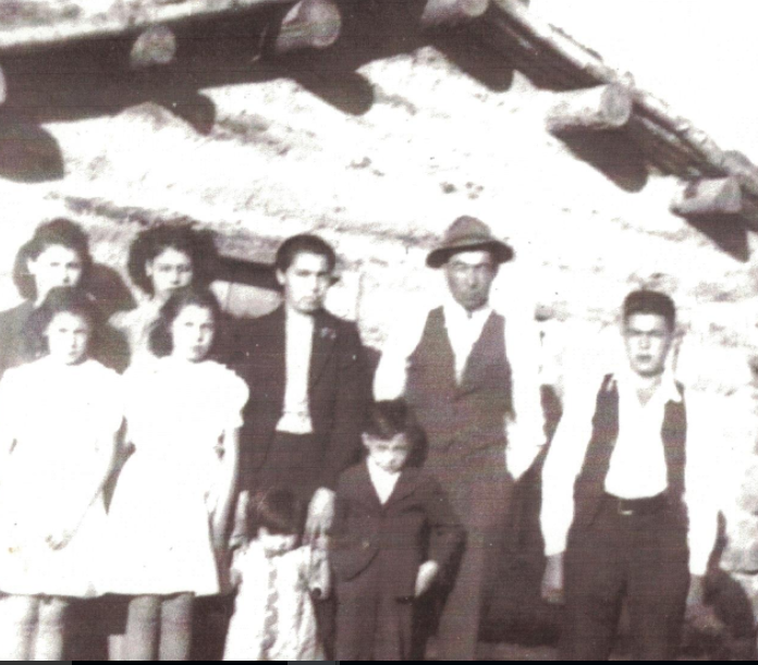 A historic photo from the mid-1940s of a family with a father and mother wearing suits, surrounded by children in front of a log house.