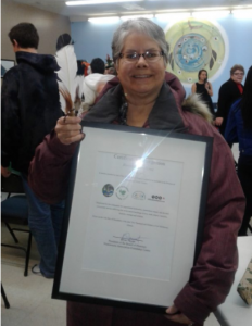 An elderly Gail Gallupe holding a certificate inside a frame, smiling looking forward with people in the background inside a hall.