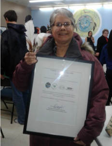 Une Gail Gallupe âgée tenant un certificat encadré, souriant et regardant vers l'avant avec des personnes en arrière-plan dans un hall.