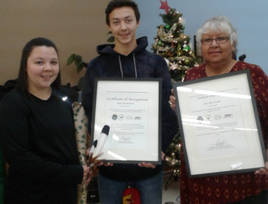 A young woman holding a feather and a young man and eldery woman holding up certificates framed in glass facing forward, a Christmas tree is on display in the background.