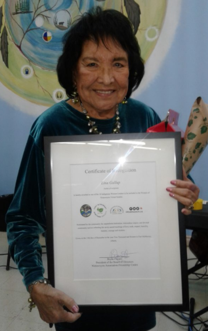 An elderly Lina Gallup stands smiling holding a large glass-framed certificate.