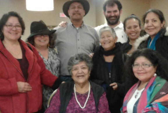 A coloured picture of a group of people, two women sitting down while the rest are standing behind them mostly everyone is smiling looking forward.