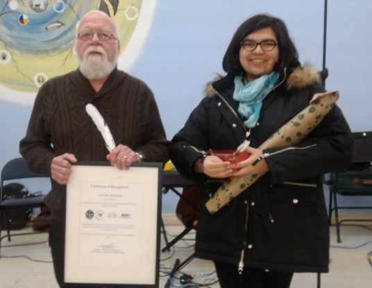 An elderly man standing beside a younger woman, the man is holding a certificate and a feather, the young woman holding a box and a bouquet of flowers.