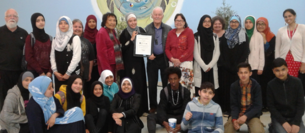 A coloured picture of group of people standing together with the elderly people standing at the back and younger ones (mostly students) sitting in front of those standing people. Most of the students are wearing hijab. In the middle is an elderly fellow named Terry Garvin and a woman beside him is wearing a hijab is holding a certificate of award. Behind Terry is the picture of the medicine wheel on the wall